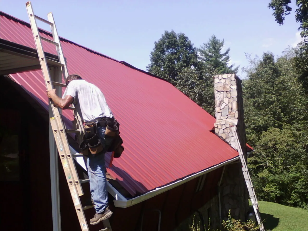 Roofer on a ladder beside a red metal roof and stone chimney — Soffit & Fascia Repair by Roofing Brothers in Pennsboro
